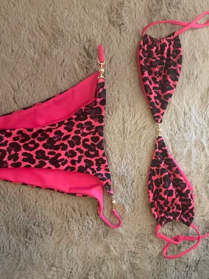Rosa leopardmönstrad bikini - Trendig bikini i rosa och svart leopardmönster med guldfärgade detaljer. Bikinin har trekantsöverdel med knyt och matchande underdel. Perfekt för sommaren och strandhäng!