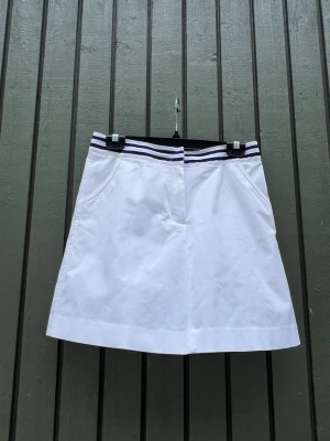 Vit minikjol/golfkjol/tenniskjol från Tommy Hilfiger - Vit minikjol från Tommy Hilfiger med sportig känsla. Kjolen har en elastisk midja med marinblå och vita ränder samt fickor fram och bak. Tillverkad i mjuk bomull och har en liten Hilfiger-logga ovanför bakfickan.