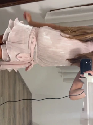 Ljusrosa omlottklänning med knyt - Säljer en ljusrosa omlottklänning med knyt i midjan. Klänningen har ett skirt, glansigt tyg och fina plisserade detaljer. Perfekt för dig som vill ha en feminin och elegant look.