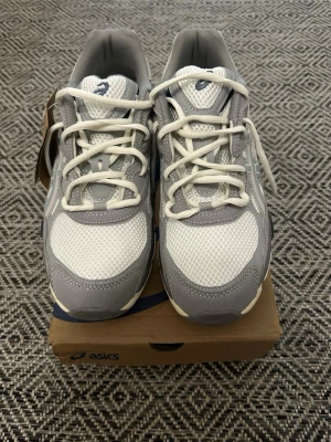 Gråvita sneakers från Asics - Snygga sneakers från Asics i grått och vitt med mesh- och syntetpaneler. Skorna har vita snören, vadderad plös och häl samt en chunky sula i beige och svart. Perfekta för dig som gillar sportig och clean stil.