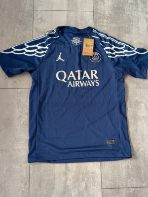 Blå PSG x Jordan matchtröja - Säljer en blå Paris Saint-Germain matchtröja från Jordan med vita detaljer och vingmönster på axlarna. Tröjan har Qatar Airways-logga på bröstet och är tillverkad i Dri-FIT-material som andas. Perfekt för fotboll eller träning.