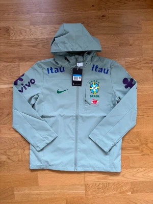 Nike Brasil vindjacka med huva - Ljusgrön vindjacka från Nike med huva och dragkedja. Jackan har Brasilien fotbollslandslagets logga på bröstet, samt tryck från sponsorer som Itaú, Vivo och Guaraná Antarctica. Tillverkad i lätt, vattenavvisande material och har meshfoder på insidan.