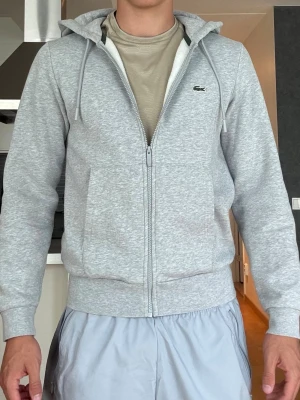 Grå Lacoste hoodie  - Säljer min gråa Lacoste zip hoodie eftersom den är för liten för mig. Den är storlek S och är i mycket bra skick. Modellen är 183.