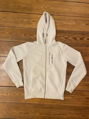 Vit hoodie från Sail Racing - Snygg vit hoodie från Sail Racing med dragkedja framtill och en extra bröstficka med zip. 