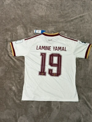 Spanien Lamine Yamal matchtröja, S, M, L finns inne   - Vit Spanien fotbollströja från Adidas med Lamine Yamal och nummer 19 på ryggen. Tröjan har vinröda detaljer, gula inslag på krage och ärmslut samt broderat landslagsmärke och Adidas-logga framtill. Korta ärmar och sportig passform, tillverkad i lätt funktionsmaterial.