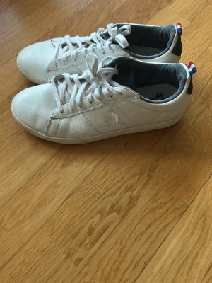 Vita Le Coq Sportif sneakers - Snygga vita sneakers från Le Coq Sportif med klassisk låg profil och vita snören. Skorna har diskret logga på sidan och en detalj i blått, vitt och rött på hälen. Ovandelen är i läder och insidan är fodrad i mörkgrått tyg.