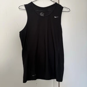 Svart Nike Dri-FIT linne - Säljer ett svart träningslinne från Nike med Dri-FIT-teknologi som håller dig torr under träningen. Linnet har en klassisk passform, rund halsringning och ett vitt Nike Swoosh-logotyp på bröstet. Perfekt för gymmet eller löprundan.