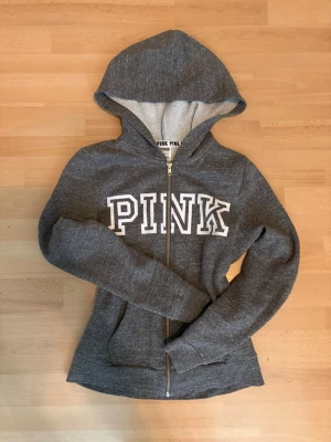 Grå zip-hoodie från PINK - Mysig grå hoodie från PINK med dragkedja framtill och stor vit logga över bröstet. Tröjan har huva med ljus insida och två fickor fram. Perfekt för en avslappnad och sportig stil. Tillverkad i mjukt sweatshirtmaterial.