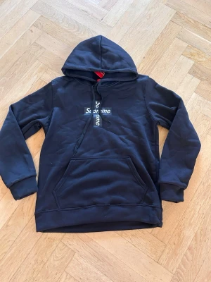 Svart hoodie från Supreme - Svart hoodie från Supreme med klassisk box logo i vitt på bröstet, både horisontellt och vertikalt. Hoodien har huva med dragsko och en stor magficka framtill. Tillverkad i mjukt bomullsmaterial, perfekt för en avslappnad streetwear-look.