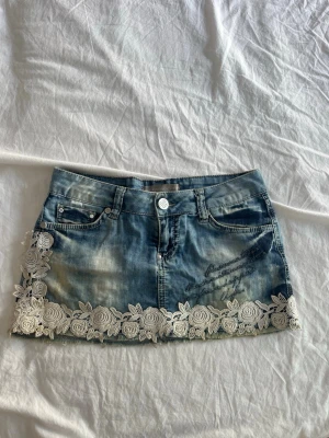 Blå jeanskjol med spetsdetaljer - Snygg blå jeanskjol med vit spets i blommönster längs nederkanten. Kjolen har klassiska jeansfickor fram och bak, samt dekorativ textbrodyr på ena sidan. Perfekt för dig som vill ha en unik och trendig look.