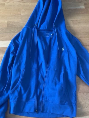 Blå zip hoodie från Polo Ralph Lauren - Snygg blå hoodie med dragkedja från Polo Ralph Lauren. Klassisk design med huva och dragsko, två fickor framtill och den ikoniska vita logon broderad på bröstet. Tillverkad i mjuk bomullsblandning som är skön att ha på sig.