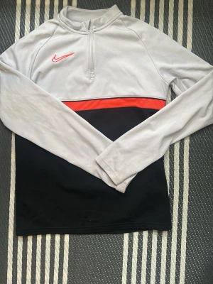 Nike Dri-Fit långärmad träningströja - Nike Dri-Fit långärmad träningströja i svart och vit med en röd detalj över bröstet. Tröjan har half zip framtill och är tillverkad i ett syntetmaterial som andas, perfekt för träning. Snygg och sportig design med klassisk Nike-logga.