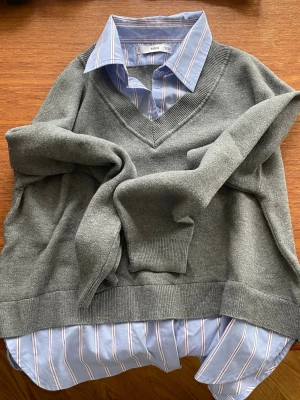 Grå V-ringad tröja från MNG - Stilren grå V-ringad tröja från MNG i mjukt stickat material. Tröjan har en fastsydd skjorta och en avslappnad passform. Enkel att matcha med olika outfits.