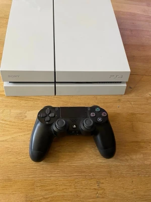Vit Sony PlayStation 4 med handkontroll - Säljer en vit PlayStation 4 från Sony tillsammans med en svart DualShock 4-handkontroll. Konsolen har en stilren, rektangulär design i plast och passar perfekt för gaming och streaming. Ikoniska PlayStation-knappar på kontrollen och modern look.