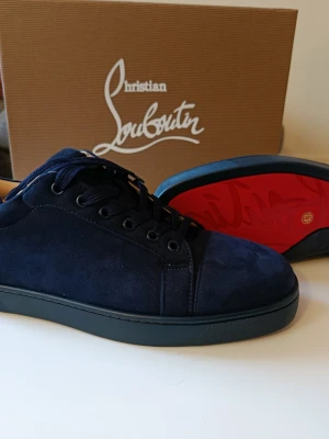 Mörkblå sneakers från Christian Louboutin - Snygga mörkblå sneakers i mocka från Christian Louboutin med klassisk röd sula och ton-i-ton broderad logga på hälen. Skorna har svarta snören och en stilren låg profil som passar perfekt till streetwear och trendiga outfits.