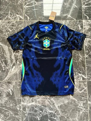 Blå Brasilien fotbollströja Jordan - Snygg blå och svart fotbollströja från Jordan i samarbete med Brasilien. Tröjan har gula detaljer, CBF-logga och Jordan-logga på bröstet. Tillverkad i ett lätt och ventilerande material, perfekt för träning eller match. Unik design med grafiskt mönster och rund hals.