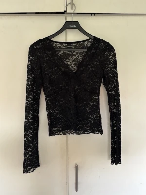 Svart spetsblus med v-ringning - Säljer en svart, långärmad blus från NA-KD i genombruten spets med v-ringad hals. Blusen är figurnära och har ett elegant, transparent utseende som passar perfekt till olika outfits.