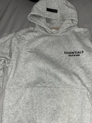 Grå Essentials Fear of God hoodie - Snygg grå hoodie från Essentials Fear of God med svart logga på bröstet. Klassisk passform med stor huva och magficka. Tillverkad i mjukt bomullsmaterial som känns riktigt skönt mot huden. Perfekt för dig som gillar streetwear och enkel stil.