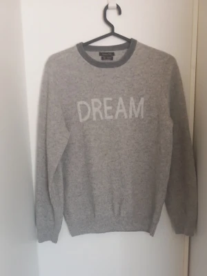 Massimo Dutti 100% ull- och kashmirtröja - Unisex grå ull- och kashmirtröja från Massimo Dutti i storlek 158–164. Tillverkad i 100 % ull/kashmir, vilket gör den mjuk och varm. Klassisk modell med rund halsringning och snyggt “DREAM”-tryck fram. Använd men i fint skick. Det finns en mindre, svagt synlig fläck på framsidan (se bild), men inga hål eller större skador. Perfekt för kalla dagar och enkel att matcha.