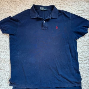 Marinblå pikétröja från Ralph Lauren - Klassisk marinblå pikétröja från Ralph Lauren med röd broderad logga på bröstet. Tröjan har korta ärmar, krage och knappar framtill. Perfekt för en avslappnad men ändå stilren look. Tecken på användning visas i bild 4 därav priset. Pris kan även diskuteras!