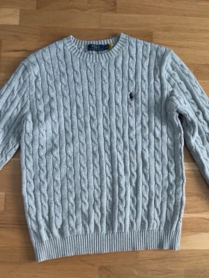 Ljusgrå kabelstickad tröja Polo Ralph Lauren - Kabelstickad tröja i ljusgrått från Polo Ralph Lauren med rund halsringning och ribbade muddar. Klassisk broderad logga på bröstet. Perfekt för lager på lager och enkel att matcha med jeans eller kjol.