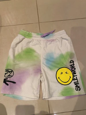 Färgglad SmileWorld pyjamas - Säljer ett par vita shorts från SmileWorld med färgstänk i grönt, lila och blått. Shortsen har tryckta smileys och textdetaljer, samt snörning i midjan. Perfekta för dig som gillar streetwear och vill sticka ut med en lekfull stil med en matchad sleeve-less hoodie.