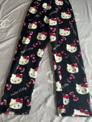 Hello Kitty mjuka loungewearbyxor - Supermysiga svarta loungewearbyxor med Hello Kitty-motiv och röda rosetter. Byxorna är i fluffigt fleece-material och har ett gulligt all-over print med vita Hello Kitty-ansikten och rosa detaljer. Perfekta för chill hemma eller sleepovers.