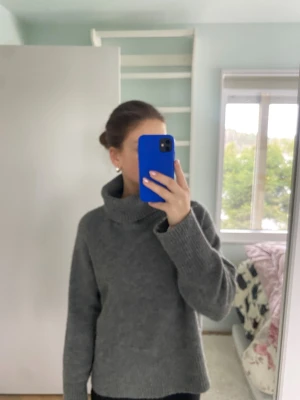 Grå polotröja i stickat material - En snygg grå stickad polotröja med bred krage och långa ärmar. Tröjan har ribbstickade muddar vid ärmslut och nederkant, och är tillverkad i ett mjukt material som passar perfekt för kyliga dagar. Enkel och stilren design som är lätt att matcha.