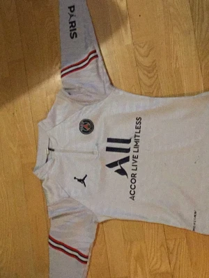 Vit långärmad PSG-tröja - Snygg vit långärmad fotbollströja från PSG med Jordan-logga och klubbmärke på bröstet. Tröjan har svarta och röda detaljer på ärmarna samt tryck med 'Accor Live Limitless' och 'Visit Rwanda'. Perfekt för dig som älskar fotboll och streetstyle.