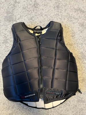 Svart säkerhetsväst från Racesafe - Säljer en svart säkerhetsväst från Racesafe med quiltad design och dragkedja framtill. Västen är vadderad och har en sportig look, perfekt för ridning. Märket syns tydligt på framsidan och västen har en bekväm passform. Den är en child’s large men går att anpassa