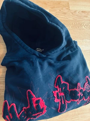 Svart hoodie från Hedi Raisér - Racer - Snygg svart hoodie från Hedi Raisér - Racer, med broderade röda och svarta detaljer framtill och på ärmen. Texten 'RACER WORLDWIDE 2021' syns i rött på ryggen. Hoodien har en stor huva och är gjord i mjukt bomullsmaterial. Perfekt för en streetwear-look. Storlek: Small
