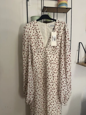 Vit långklänning med körsbär från Adoore - Vit långklänning från Adoore med rött körsbärsmönster, v-ringning och långa puffärmar. Klänningen har en lätt texturerad yta och stängs med dragkedja i ryggen. Perfekt för dig som gillar romantiska detaljer och en feminin siluett.