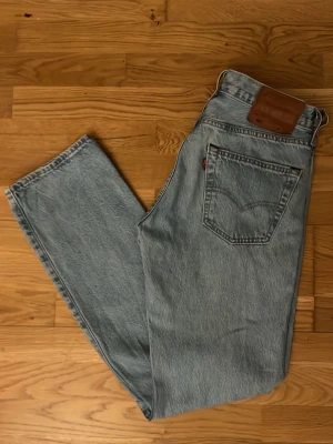 Levi's 501 - Säljer ett par klassiska ljusblå Levi's Premium jeans med rak passform och hög midja. Jeansen har fem fickor, bruna sömmar och en ikonisk läderpatch bak i midjan. Perfekta för en avslappnad och tidlös stil.