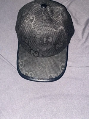 Svart keps från Gucci med GG-mönster - Snygg svart keps från Gucci med det klassiska GG-mönstret över hela kepsen. Kepsen har böjd skärm med svart kant i läderimitation och justerbar rem bak. Tillverkad i slitstarkt tyg med detaljer i svart. Perfekt för dig som vill ha en exklusiv och trendig look.