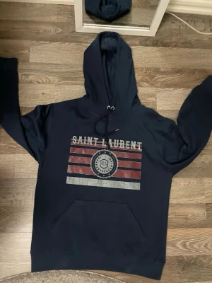 Mörkblå Saint Laurent hoodie - Mörkblå hoodie från Saint Laurent med stor huva och magficka. Framsidan har ett tryck med texten 'SAINT LAURENT' och röda, vita och gråa ränder samt ett emblem i mitten. Tillverkad i mjukt bomullsmaterial och har ribbade muddar vid ärmslut och nederkant.