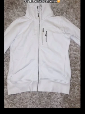 Vit Sail Racing hoodie med dragkedja - Snygg vit hoodie från Sail Racing med hel dragkedja framtill och hög krage. Har en bröstficka med dragkedja och Sail Racing-logga, samt två sidofickor. Perfekt för en clean och sportig look. Tillverkad i mjukt material för extra komfort.