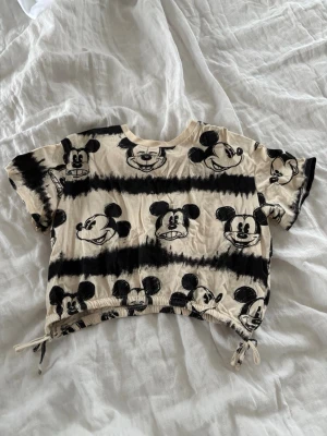 Beige croppad topp med Mickey Mouse - Croppad topp i beige med svart Mickey Mouse-tryck och batikinspirerade ränder.😍 Toppen har korta ärmar och snörning i nederkanten på båda sidor för justerbar passform. Perfekt för en avslappnad och lekfull stil.💓💓