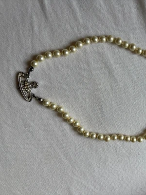 Vivienne Westwood pärlhalsband - Ikoniskt halsband från Vivienne Westwood med vita pärlor och det klassiska Orb-hänget i silverfärg med gnistrande stenar. Perfekt statement-piece för dig som gillar unika accessoarer och vill sticka ut lite extra.