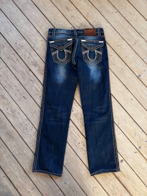 True Religion Y2K distressed washed jeans - Ett par True Religion jeans i mörkblå denim med kontrasterande sömmar i beige och vitt.   Jeansen har en rak passform och detaljerade fickor bak med True Religion's signaturhästskodesign.   Midjan är normalhög och byxorna har en klassisk knapp- och dragkedjegylf. Innerfodret i fickorna har ett mönster med röda gitarrer och True Religion-logotyper.  Mått: Midja 39 cm Längd 102 cm