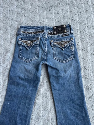 Miss Me jeans med paljettdetaljer - Snygga blå jeans från Miss Me med utsvängda ben och coola paljettdetaljer på bakfickorna. Jeansens bakfickor har även dekorativa knappar och broderad logga. Jeansen är ny skick, säljer dom pga att dom inte passar mig, andvända 1 gång❣️