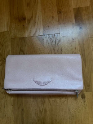 Ljusrosa kuvertväska från Zadig & Voltaire - Snygg ljusrosa väska från Zadig & Voltaire. Väskan har silverfärgad kedjerem och ett innerfack samt ett på baksidan och i mitten delen av väskan. Köpt på zalando för ett tag sen men är i nyskick och knappt använd. Skriv för mer frågor eller bilder vid intresse. 