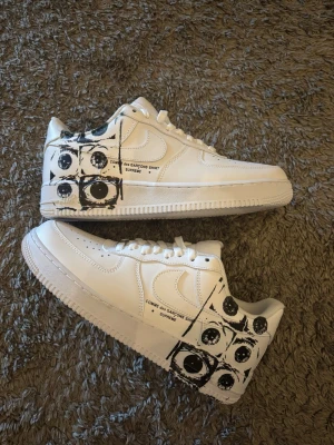 Nike x Comme des Garçons x Supreme sneakers - Unika vita sneakers från Nike i samarbete med Comme des Garçons Shirt och Supreme. Skorna har svart grafiskt ögonmönster på hälen och sidan, samt texttryck. Tillverkade i läder med klassisk låg siluett och vita snören. Perfekta för dig som gillar streetwear och statement pieces.