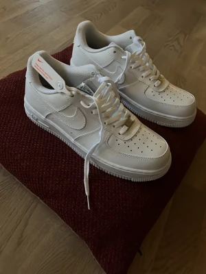Nike Air Force 1 vita sneakers - Klassiska vita Nike Air Force 1 sneakers med låg profil och rund tå. Skorna har snörning, perforerade detaljer på tån och är tillverkade i skinn med syntetdetaljer. Swoosh-logga på sidan och diskret AIR-text på sulan. Perfekta för en clean och stilren look. Använda 3 gånger och på vänster sko är sulan lite skadad men syns ju inte när man har på sig skorna.