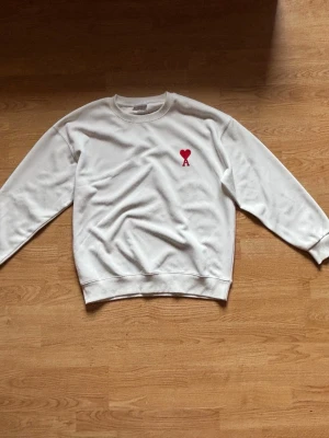 Vit ami paris sweatshirt med röd hjärtlogo - Vit sweatshirt från AMI Paris med rund hals och lång ärm. På bröstet finns en röd broderad hjärtlogo med A. Tröjan har ribbade muddar vid ärmslut och nederkant. Perfekt för en clean och stilren look.