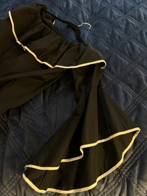 Svart offshoulder volangblus - Trendig svart offshoulder-blus med stora volangärmar och vita kontrastkanter. Blusen har en lös passform och är perfekt för dig som vill sticka ut med en cool och unik topp. Materialet känns lätt och luftigt, och volangdetaljerna ger en extra touch.