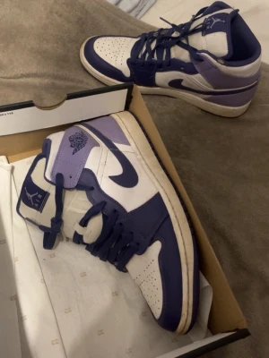 Air Jordan 1s mid - Säljer ett par Air Jordan 1 High sneakers i lila och vitt. Skorna har klassisk hög siluett, Nike swoosh på sidorna och Jordan Wings-logga vid ankeln. Ovandelen är i läder med perforerad tåbox för extra ventilation. Perfekta för dig som vill sticka ut med din stil. Dom är knappast använda, eftersom jag bara använt ett annat par 