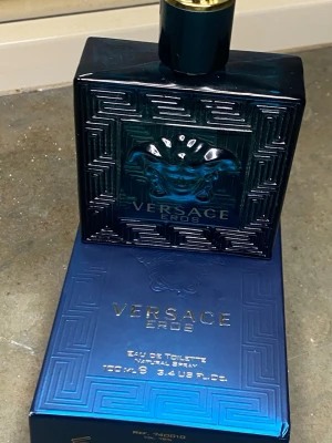 Versace Eros Eau de Toilette 100ml - Versace Eros Eau de Toilette i en lyxig, fyrkantig flaska i mörkblått glas med Medusa-huvud i relief och gulddetaljer. Flaskan rymmer 100 ml och kommer i en matchande blå förpackning med guldtext. En ikonisk doft för dig som gillar exklusiva parfymer.