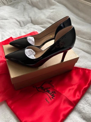 Svarta lackpumps från Louboutin - Säljer ett par klassiska svarta pumps i lack från Christian Louboutin. Skorna har spetsig tå, hög klack och den ikoniska röda sulan. Tillverkade i glansigt läder med beige innersula och elegant siluett. Perfekta för dig som vill ha en tidlös och lyxig look.
