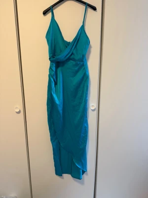 Turkos långklänning med omlott - Säljer en turkos långklänning med smala axelband och omlott-detalj framtill. Klänningen har en elegant v-ringning och ett draperat fall som ger en snygg siluett. Perfekt för dig som vill sticka ut med färg! 💙