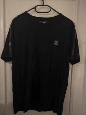 Svart t-shirt från Plein Sport - Säljer en svart t-shirt från Plein Sport med tryck på ärmarna och en liten logga på bröstet. Klassisk rund hals och normal passform. Tillverkad i mjuk bomull som känns skön mot huden. Perfekt för dig som gillar stilrena och sportiga plagg.